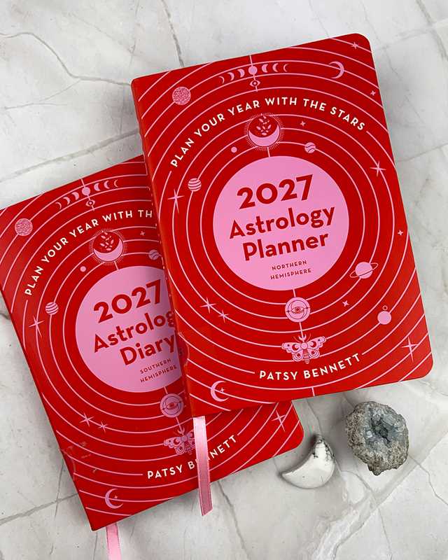 Two red 2027 Astrology Planner notebooks by Patsy Bennett are arranged on a light marble surface with small decorative props, including a white moon and a gray geode.