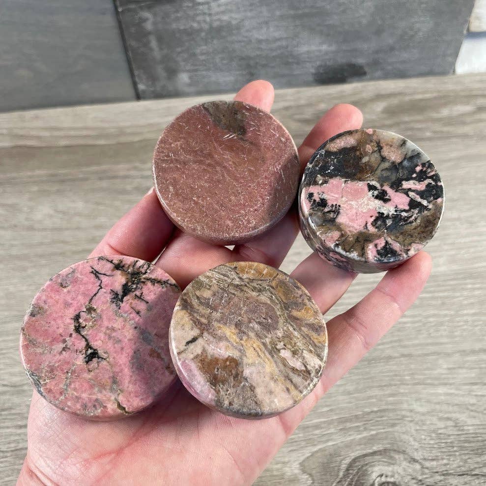 Four round, Rhodonite stones with marbled patterns held in a hand on a wooden surface.