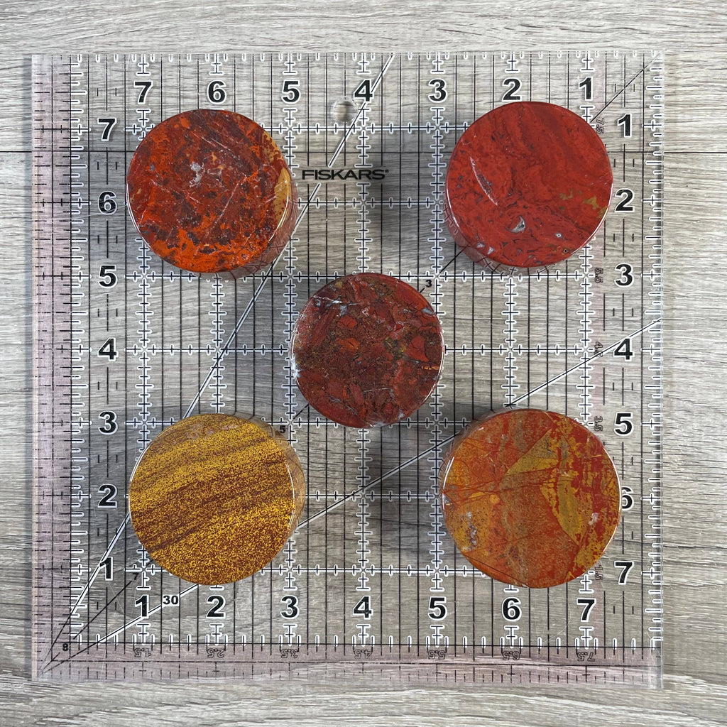 Five red jasper disks arranged on a clear Fiskars 8x10 inch quilting ruler over a gray wood-grain surface.
