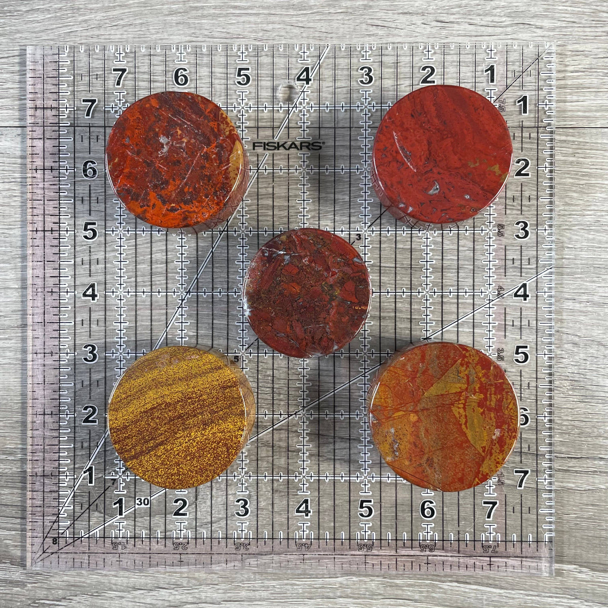 Five red jasper disks arranged on a clear Fiskars 8x10 inch quilting ruler over a gray wood-grain surface.