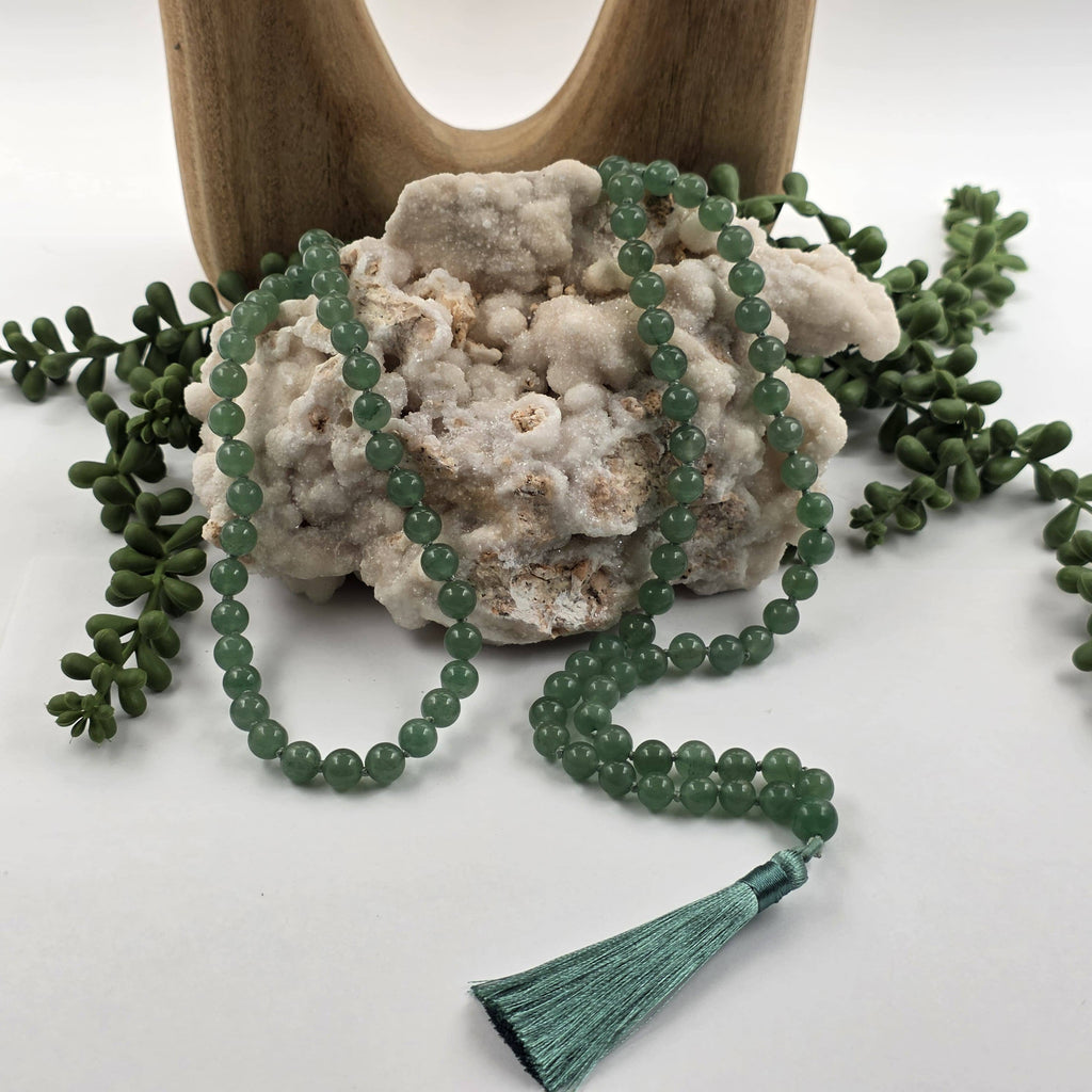 A green beaded mala prayer necklace with a matching tassel, draped over a white textured stone with small green leaves around it and a wooden stand in the background.