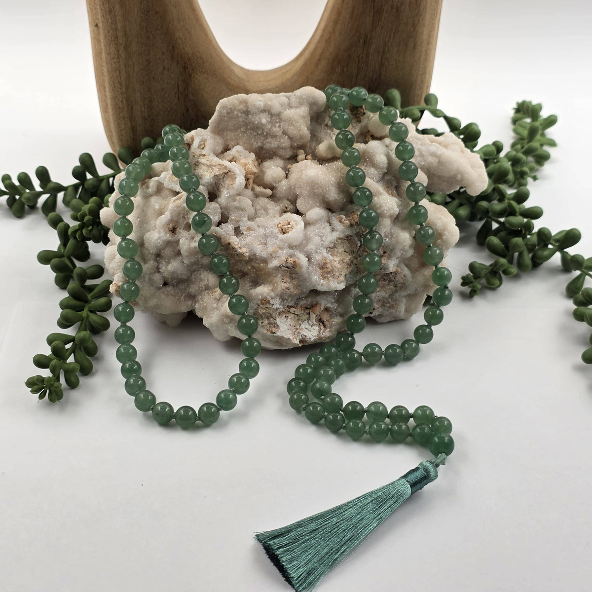 A green beaded mala prayer necklace with a matching tassel, draped over a white textured stone with small green leaves around it and a wooden stand in the background.