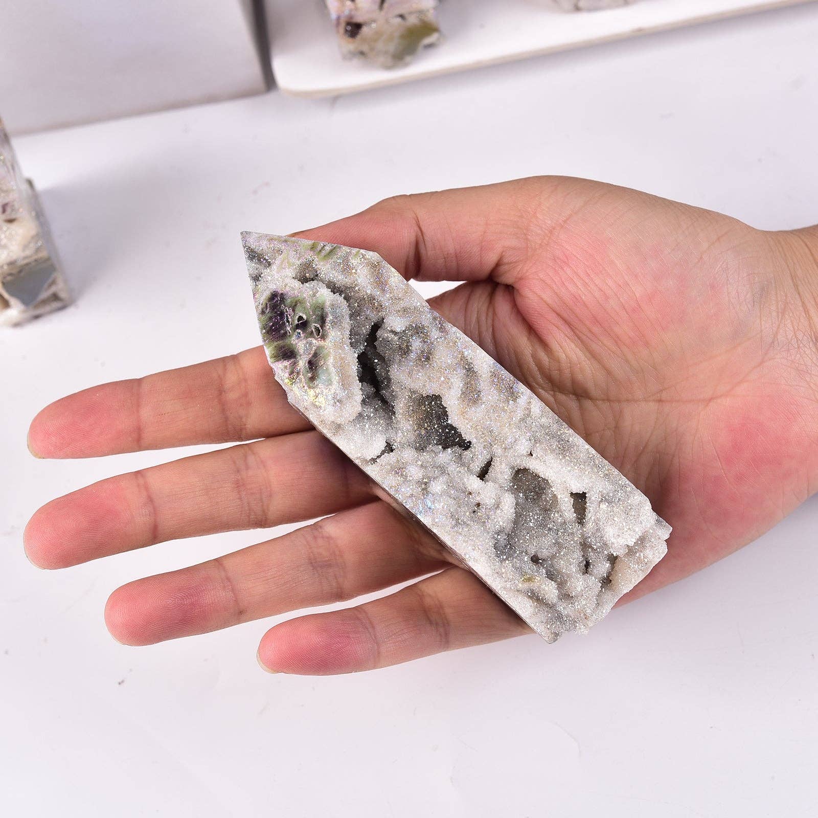 A hand holding a pointed crystal cluster specimen with a rough, textured white and gray surface showing dark inclusions, photographed against a white tabletop.