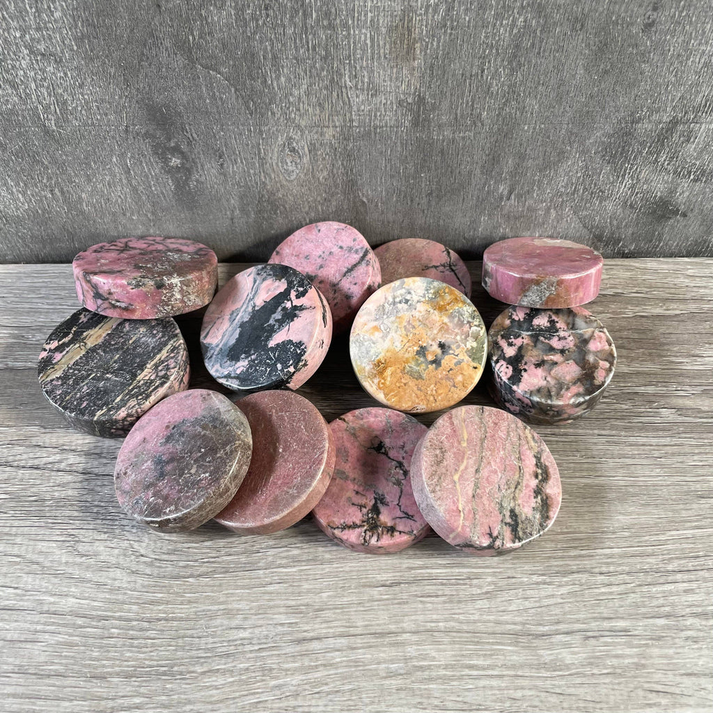 A group of round, polished rhodonite disks, arranged on a gray wood-textured surface against a gray concrete wall.