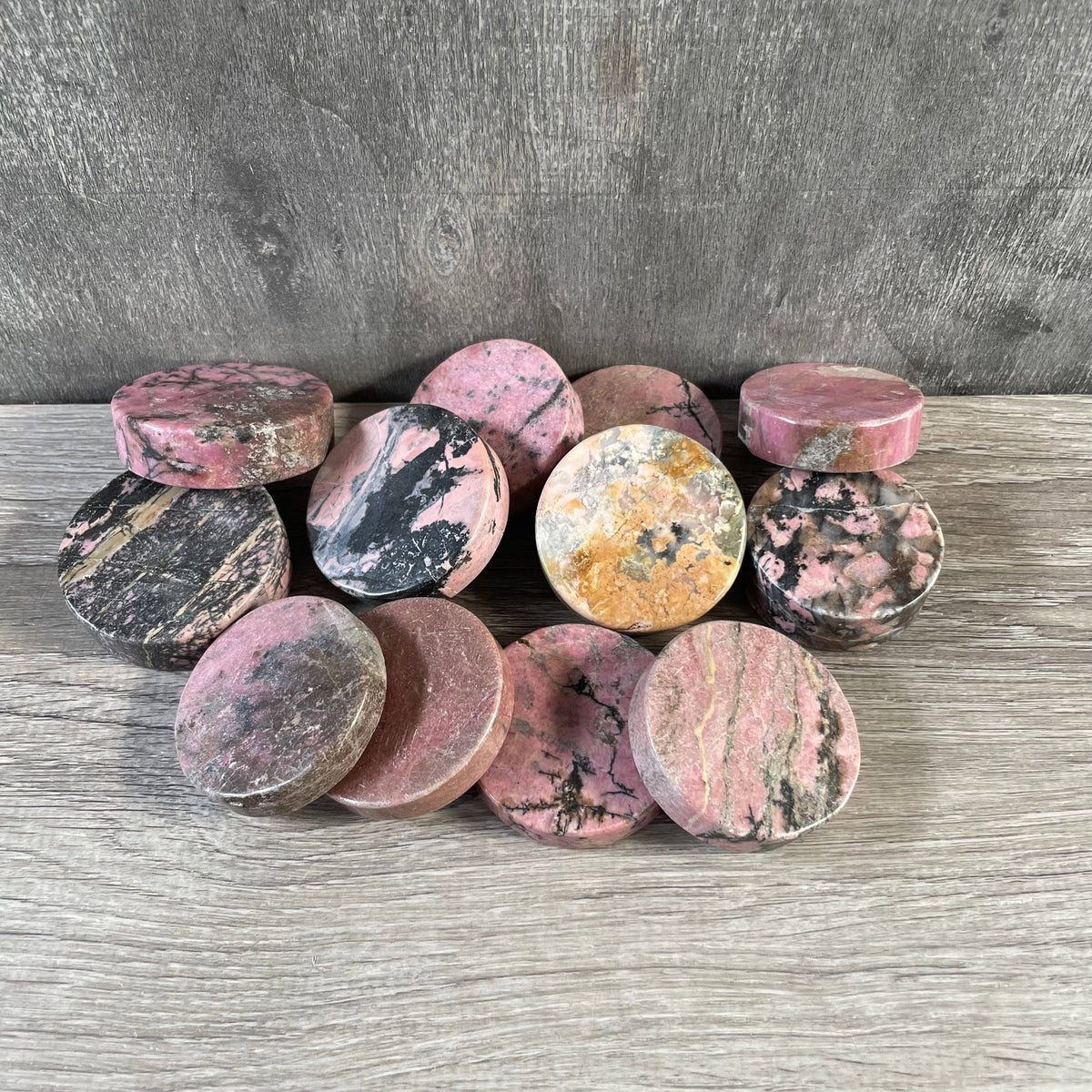 A group of round, polished rhodonite disks, arranged on a gray wood-textured surface against a gray concrete wall.