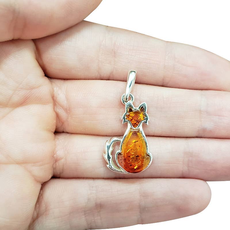 A silver pendant shaped like a sitting cat made of amber, shown resting on a person's palm to demonstrate scale.