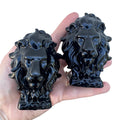 Two glossyBlack Obsidian Lion Head sculptures held in a person's hand against a light gray wall.