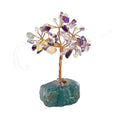 A fluorite crystal tree figurine featuring a twisted brown wire trunk and branches adorned with small purple, clear, and pale yellow amethyst stones, mounted on a dark green raw mineral base, shown against a plain white background. 63 beads