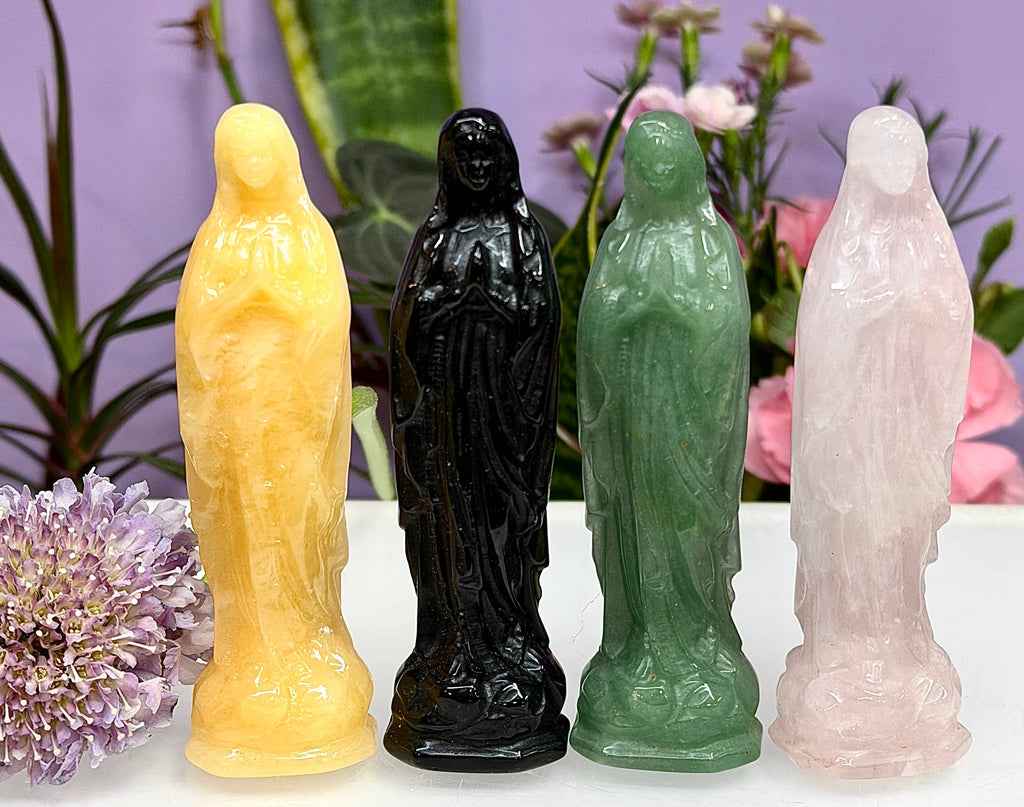 Four small standing figurines carved from colored stone, shown side by side on a white surface with flowers and greenery in the background. From left to right, the figures are yellow, glossy black, green, and pale pink.