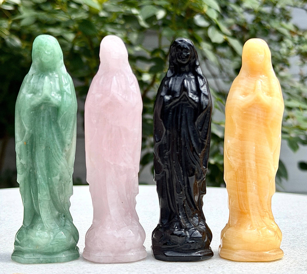 Four small carved figurines of a robed figure standing side by side in different stone colors: mint green, pale pink, glossy black, and warm yellow, photographed outdoors on a light surface with blurred greenery behind.