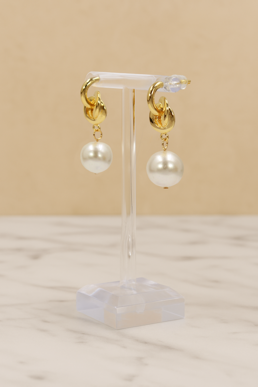 Gold hoop earrings with white pearls on a clear stand against a beige background