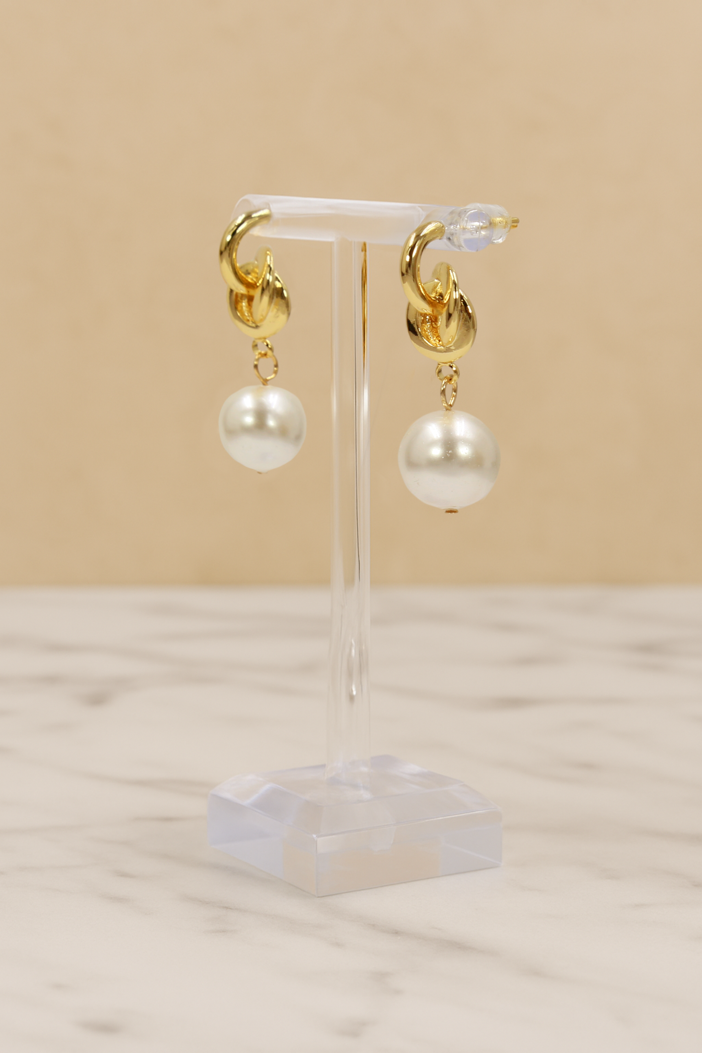 Gold hoop earrings with white pearls on a clear stand against a beige background