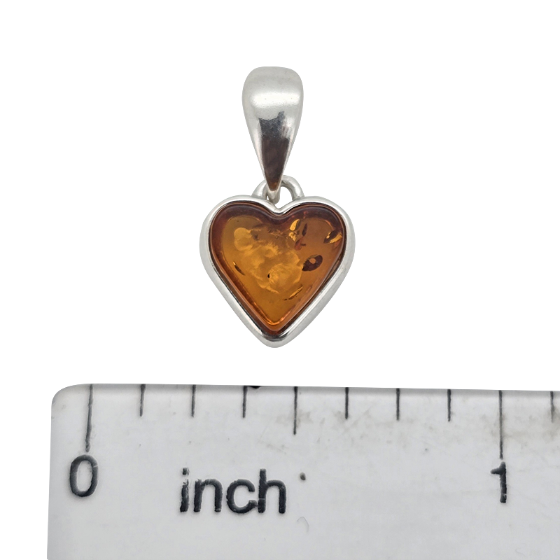 A heart-shaped amber pendant in a silver bezel setting with a bail, shown above a white inch ruler for scale on a white background.