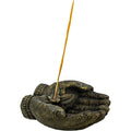 A dark textured sculpture of two hands holding a thin gold incense stick, set against a solid white background.