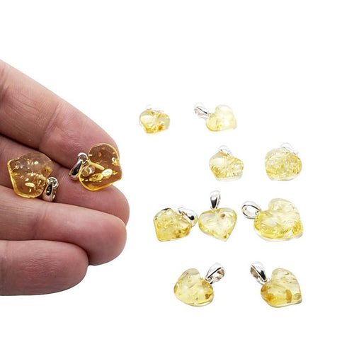 Several small translucent yellow amber heart-shaped gemstone pendants with silver bail loops are shown on a white background, with a hand holding two of them for scale on the left.