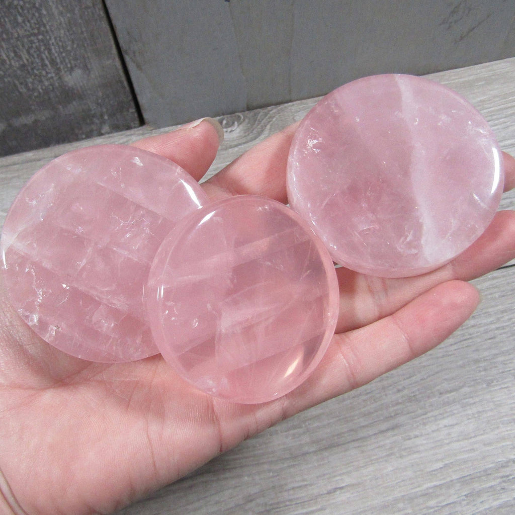 Three round pink rose quartz crystal disks held in a person's hand against a gray wood-grain background.