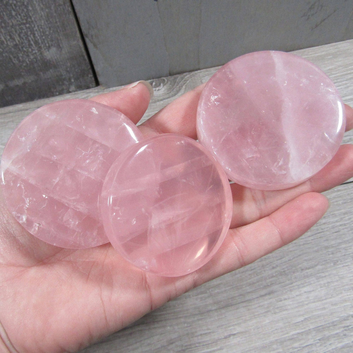 Three round pink rose quartz crystal disks held in a person's hand against a gray wood-grain background.