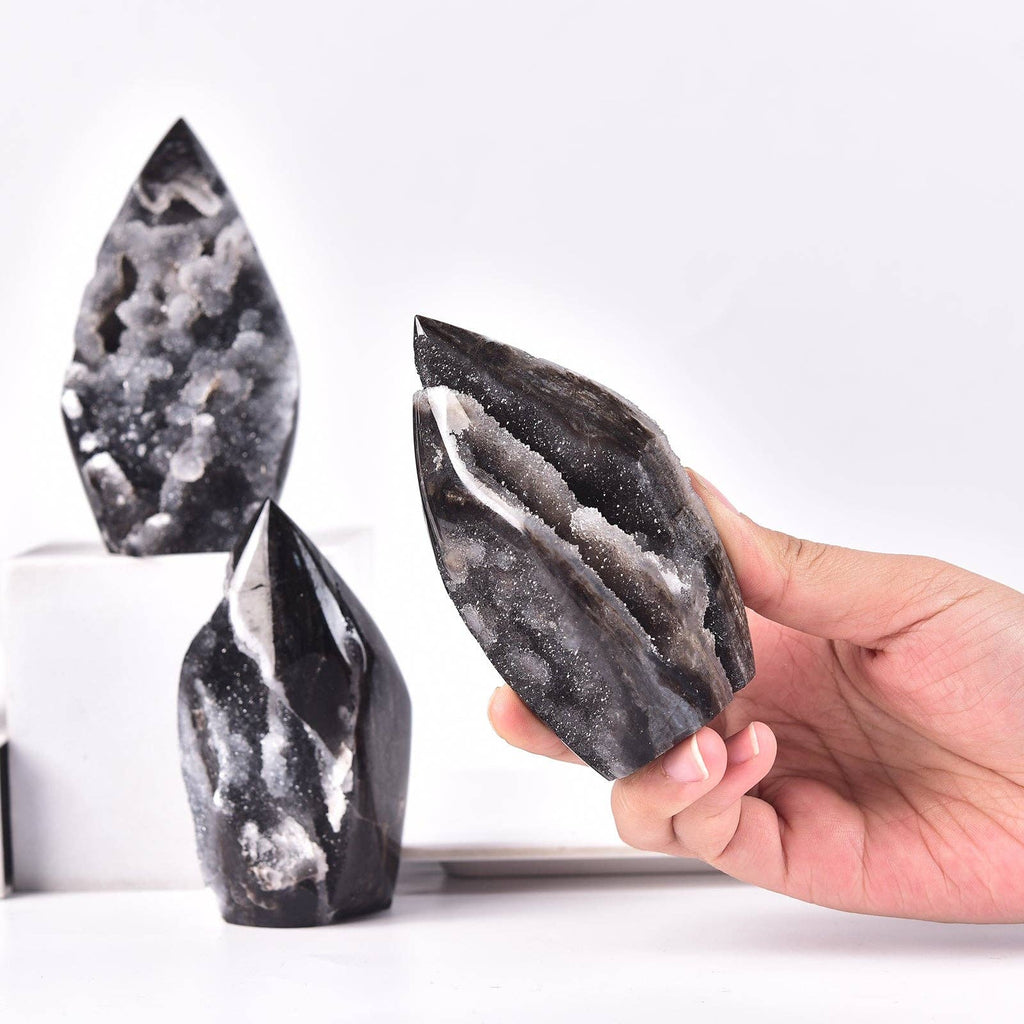 A hand holds a polished black and white geode crystal point, with two more similar crystals displayed in the background on a clean white surface.
