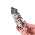 A hand holding a pointed crystal cluster with a mottled pattern of gray, white, black, and subtle iridescent purple tones against a plain white background.