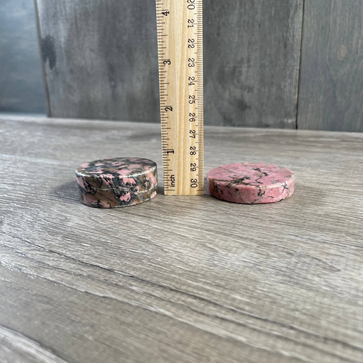 Two rhodonite stones on a wooden surface with a ruler for scale showing varying thicknesses.